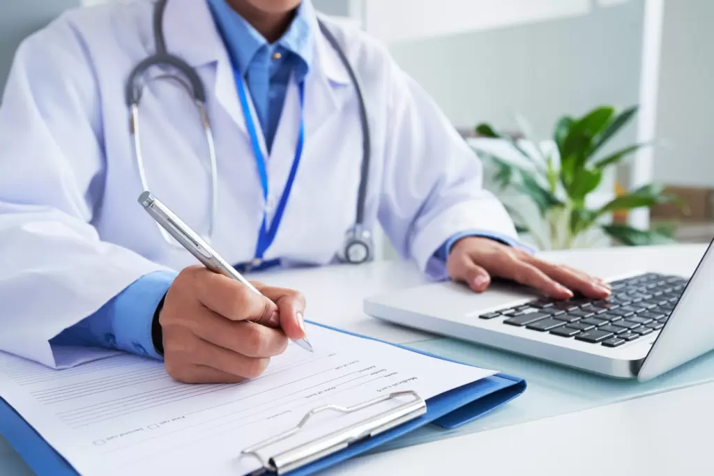 The image captures a close-up of a healthcare professional's hands as they are engaging in dual tasks: writing on a medical form with a pen and operating a laptop keyboard. The individual is wearing a white lab coat with a stethoscope draped around the neck and a light blue shirt underneath. On the desk, there is a clipboard with a document attached to it, and a laptop computer directly in front of the person. The background is soft and bright, with indoor plants adding a touch of greenery, suggesting a clean and serene medical office environment. This image is apt for a section discussing the implementation of a full-funnel marketing strategy and mapping the patient journey, highlighting the meticulous work healthcare providers put into managing patient information and care plans.