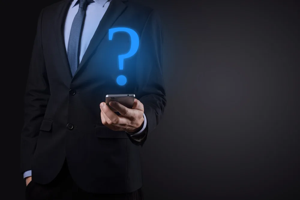 A conceptual representation of a businessman with a glowing blue question mark hologram in place of his head, symbolizing the inquiry into AI technologies, holding a smartphone in a dark suit against a black backdrop.