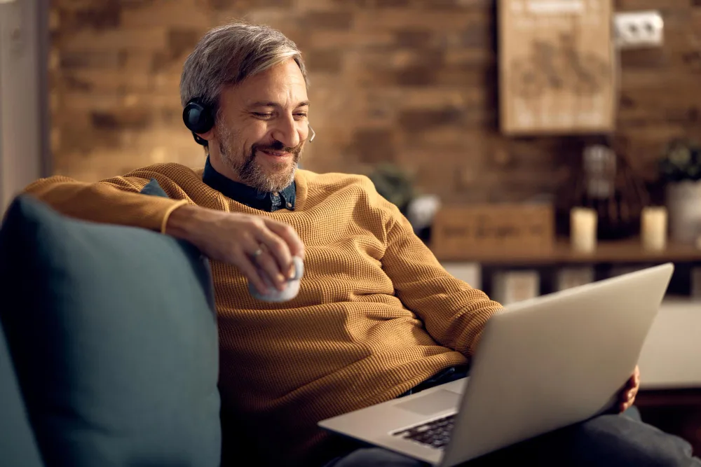 Contented man in a yellow sweater using a laptop at home, symbolizing the relaxed integration of AI in everyday work and SEO strategies.