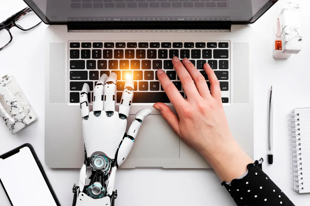 A human hand and a robot hand collaborating on a laptop, highlighting the partnership between humans and generative AI in the workplace.