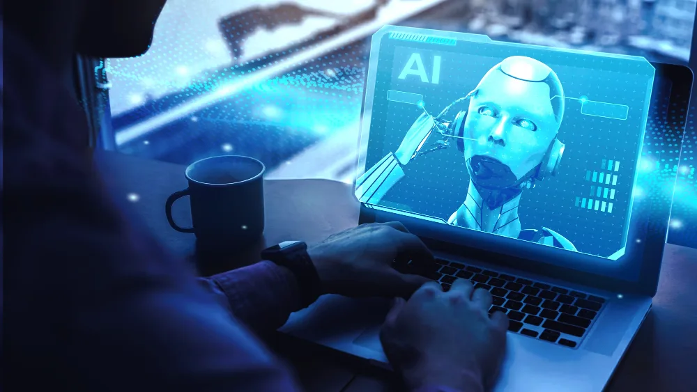 A close-up view of an individual's hands typing on a laptop displaying a 3D model of a humanoid robot's head with the label 'AI'. The screen casts a blue glow, and there is a coffee mug on the table, suggesting a work environment focused on artificial intelligence development.