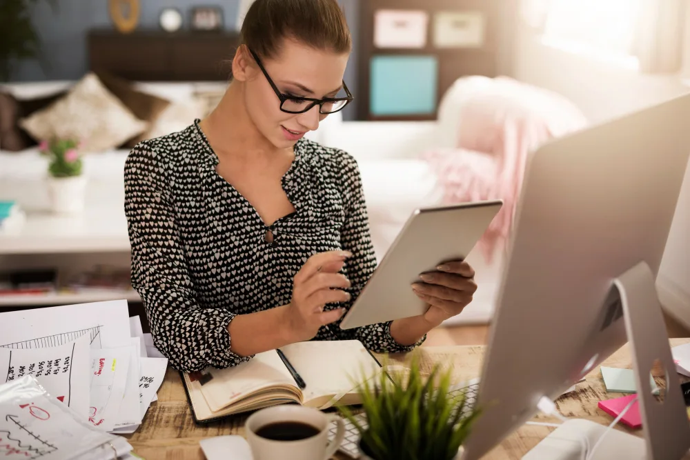 A content strategist focused on her work in a well-lit home office. She's wearing glasses and a patterned blouse, intently using a tablet while surrounded by charts, a notebook, a cup of coffee, and a desktop computer