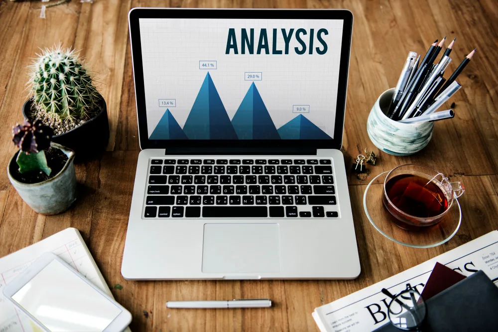 A laptop on a wooden desk displays a bar graph titled "ANALYSIS" with various percentages, indicating a focus on performance analysis for content marketing. Accompanying the laptop are office essentials: a smartphone, a pen, potted cacti, a cup of tea, and a ceramic holder with pencils, suggesting an organized and productive workspace. There's also a pair of glasses and some reading material, which includes a section titled "Business," hinting at a professional environment.