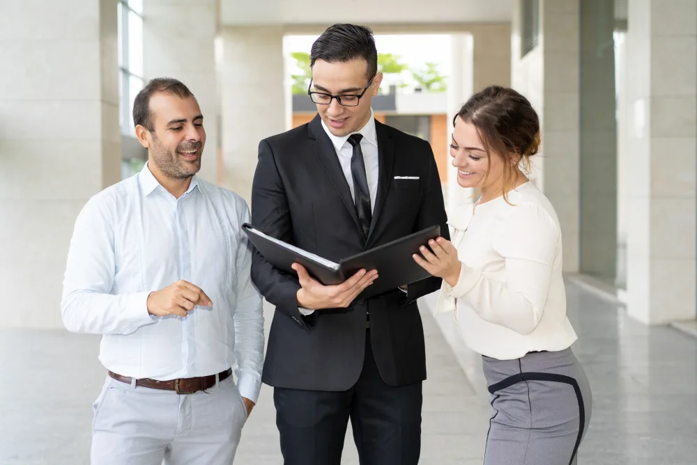 Three business professionals collaboratively reviewing a document in a portfolio. The scene is set in a bright, modern corridor, with the focus on a man in a black suit holding the portfolio. A woman in a white blouse and a man in a white shirt, both appearing engaged and pleased, are looking on. The interaction suggests a positive dialogue, likely about performance analysis or business results.