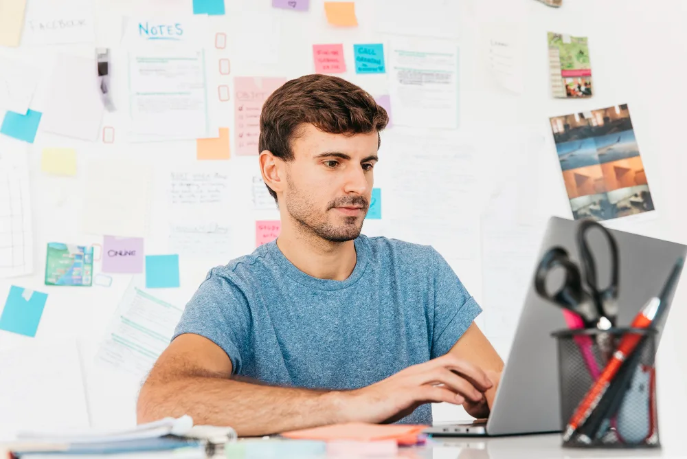 A focused male professional working on a laptop in a creative workspace adorned with a variety of sticky notes, papers, and pictures pinned to the wall behind him. The environment suggests a brainstorming session, likely for strategies to enhance audience engagement in a B2B setting, possibly utilizing AI technology to optimize interactions.