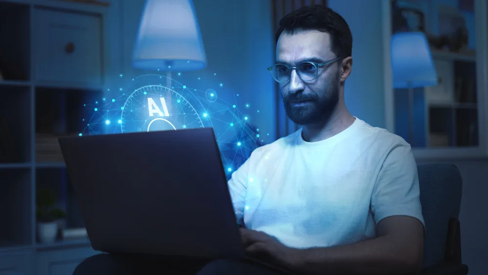 A man with glasses is focused on his laptop in a dimly lit room illuminated by the blue glow from the screen, which displays a holographic "AI" symbol, representing artificial intelligence. This setting suggests that he might be using AI-powered article writing services or software for content creation or analysis. The ambiance is serene and technology-driven, emphasizing the blend of human interaction with advanced AI capabilities.