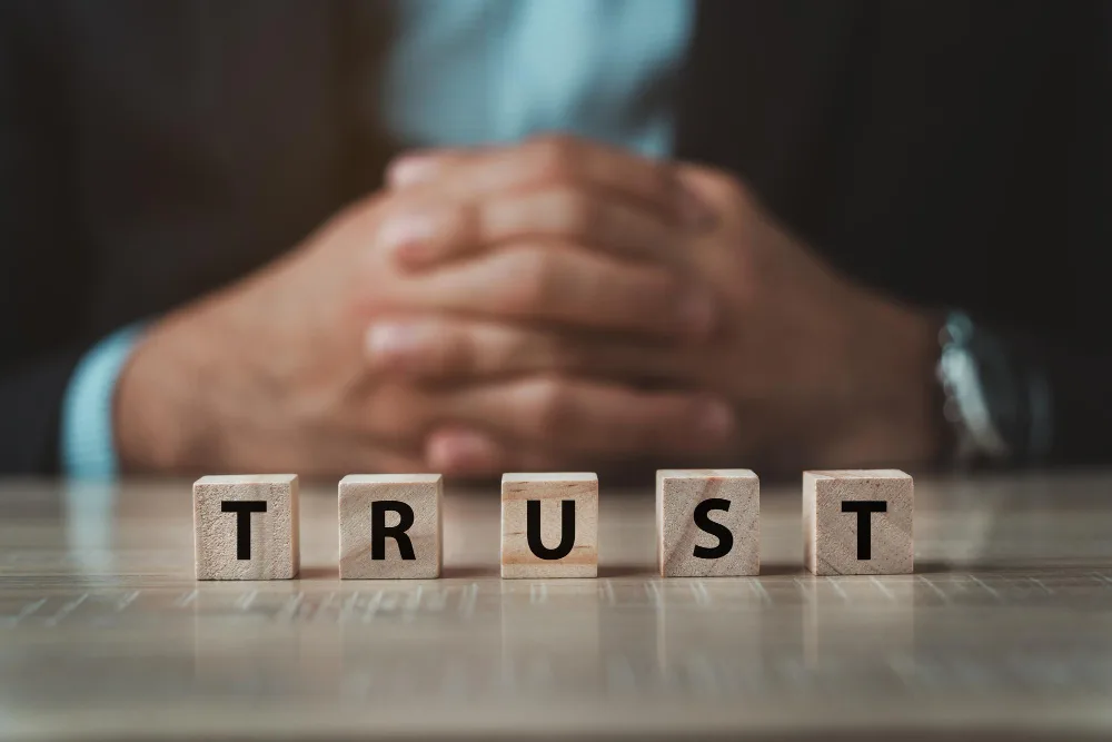 The word "TRUST" spelled out in wooden block letters on a table with a person in business attire sitting behind it, their hands clasped together, symbolizing a professional commitment to trustworthiness and reliability in business dealings.