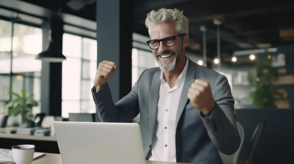 A jubilant middle-aged man with grey hair, wearing glasses, a grey suit, and a white shirt, is seated in a modern office environment. He's raising his fists in a victorious gesture as he looks at his laptop screen, possibly indicating a successful outcome, such as receiving positive feedback on article writing services or securing a new client through his content marketing efforts. The backdrop of the office includes indoor plants and large windows, contributing to an airy and bright workspace.