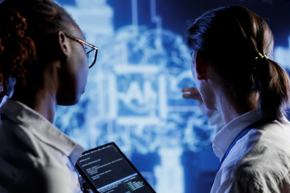 Two professionals are engaged in a technological task with one holding a tablet displaying code. Behind them is a digital screen with complex blueprints, illustrating a high-tech environment indicative of cognitive computing and AI development.