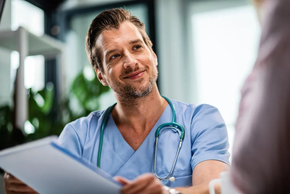 A male healthcare worker in blue scrubs, looking up and smiling, potentially discussing patient care strategies involving AI technology, suggesting a collaborative AI future in healthcare.