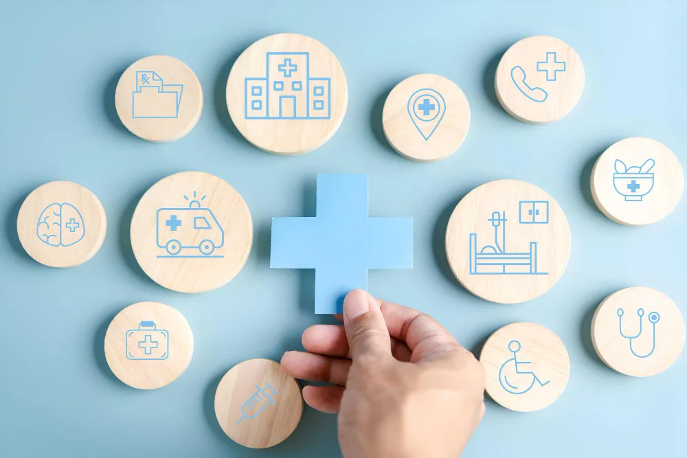 A hand positioning a blue cross symbol among wooden tokens engraved with various healthcare and medical icons on a light blue background. This could represent the importance of accessible healthcare services and the comprehensive nature of healthcare offerings. It symbolizes organization, connectivity, and the central role of health in our lives. The image is simple yet powerful in conveying the message of integrated and holistic healthcare.