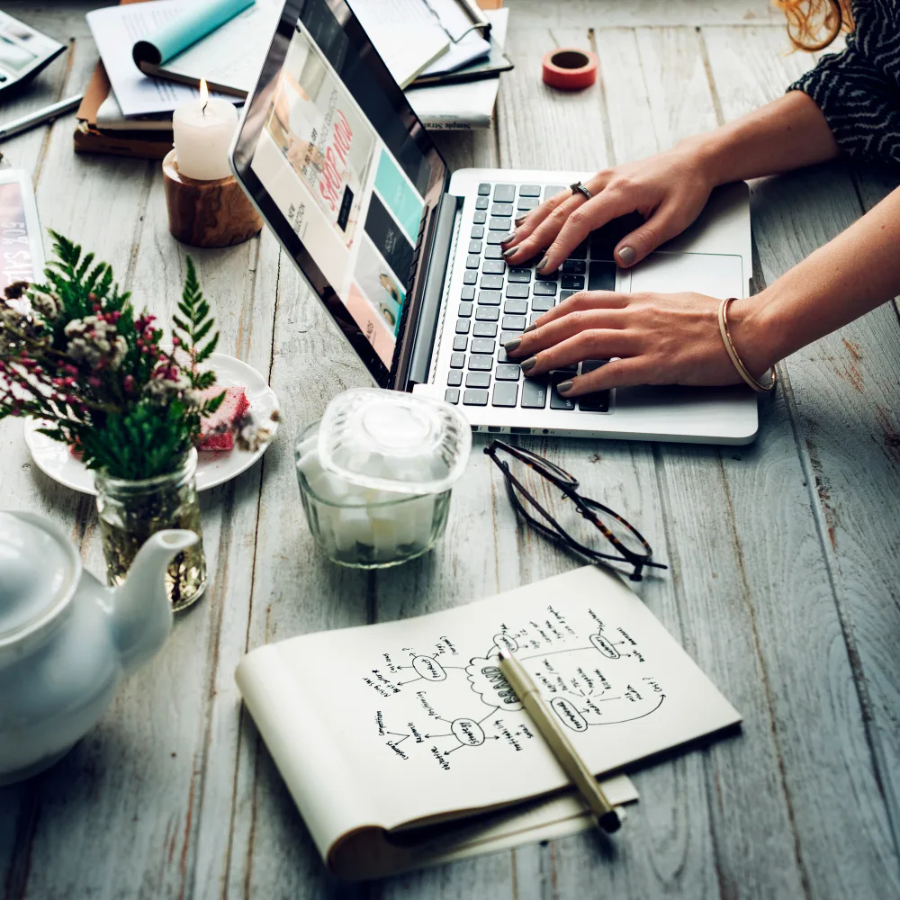 An individual is using a laptop at a wooden desk strewn with creative tools and a notepad with drawn diagrams, illustrating a scene of content optimization rooted in deep industry expertise.