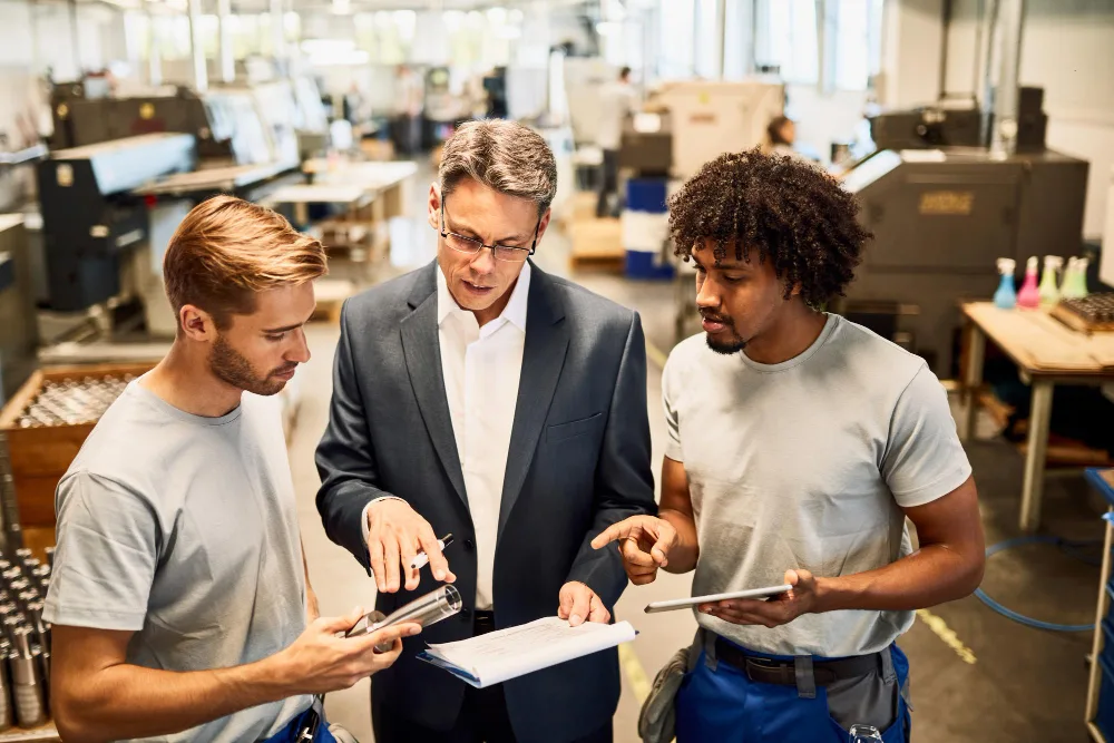 Three industry professionals are collaboratively working in a factory setting, with one holding a smartphone and another referencing a tablet, showcasing their industry expertise as they optimize content for a specific project.