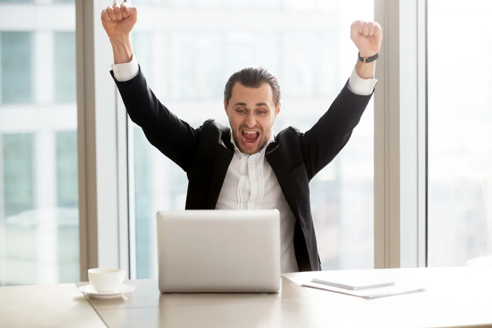 Exuberant businessman with arms raised in a victory gesture in front of a laptop in an office, symbolizing the success of an industry expert or thought leader.