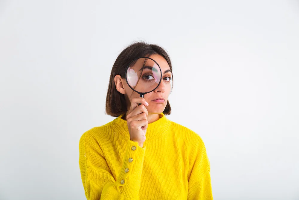 A woman peers through a magnifying glass, her eye enlarged in focus, representing the scrutiny and detailed examination necessary when implementing advanced techniques in marketing innovation.