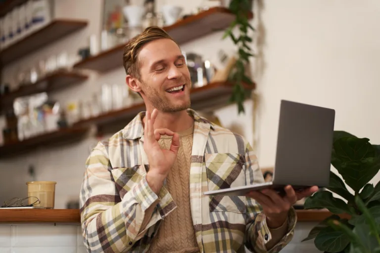 A man in a café gives a positive 'OK' sign during an online interaction, potentially discussing post-AI content strategies